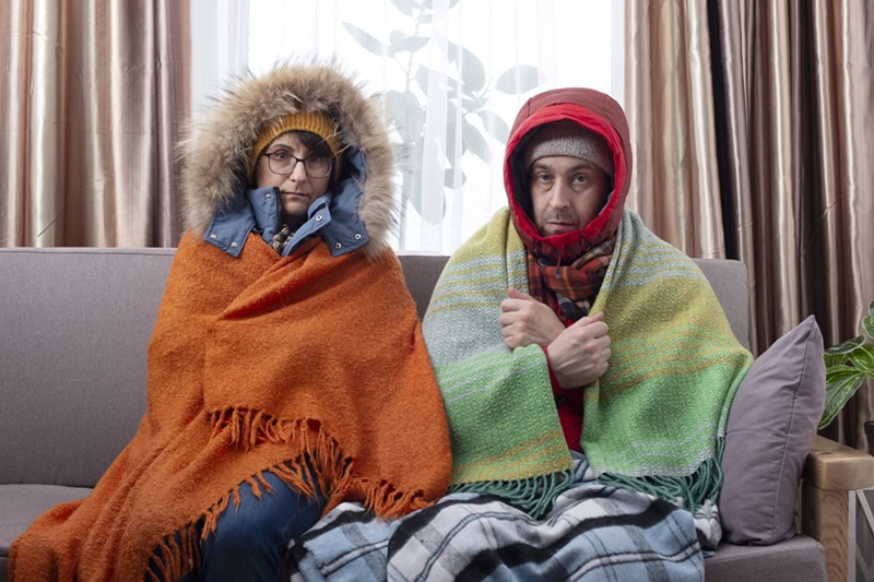 A man and a woman sitting side by side on a sofa indoors, wrapped in heavy winter coats, hoods, and blankets while looking cold and uncomfortable.