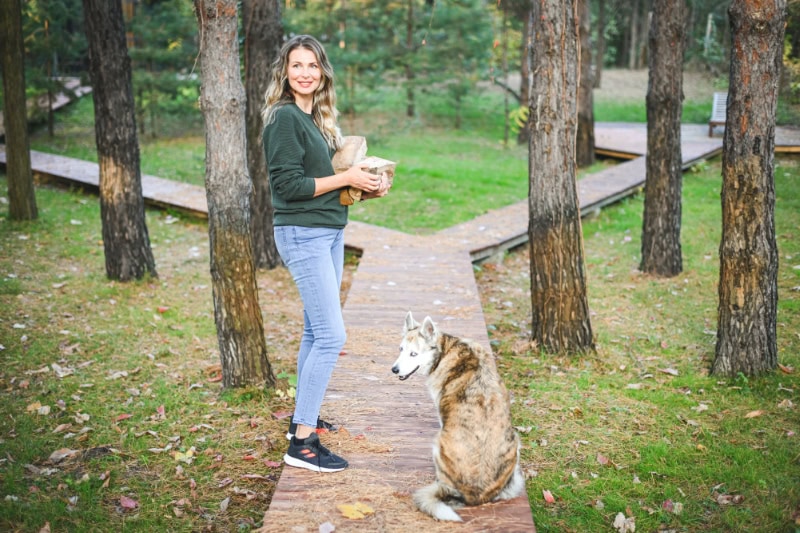 A woman carrying logs of wood stands along a wooden path in a forest setting with a dog sitting nearby.