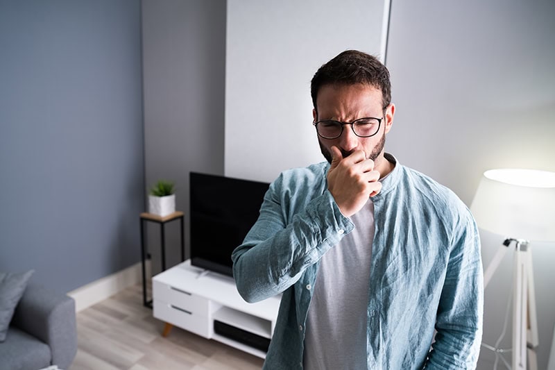 A man with glasses standing in a living room holds his hand up to cover his nose or mouth, looking uncomfortably bothered by a bad smell or feeling unwell.